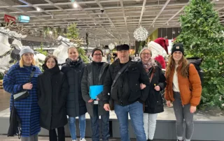 Sieben Personen in Winterkleidung stehen in einem Innenraum in der Nähe von Weihnachtsschmuck, darunter eine große Weihnachtsmannfigur und ein künstlicher Baum.