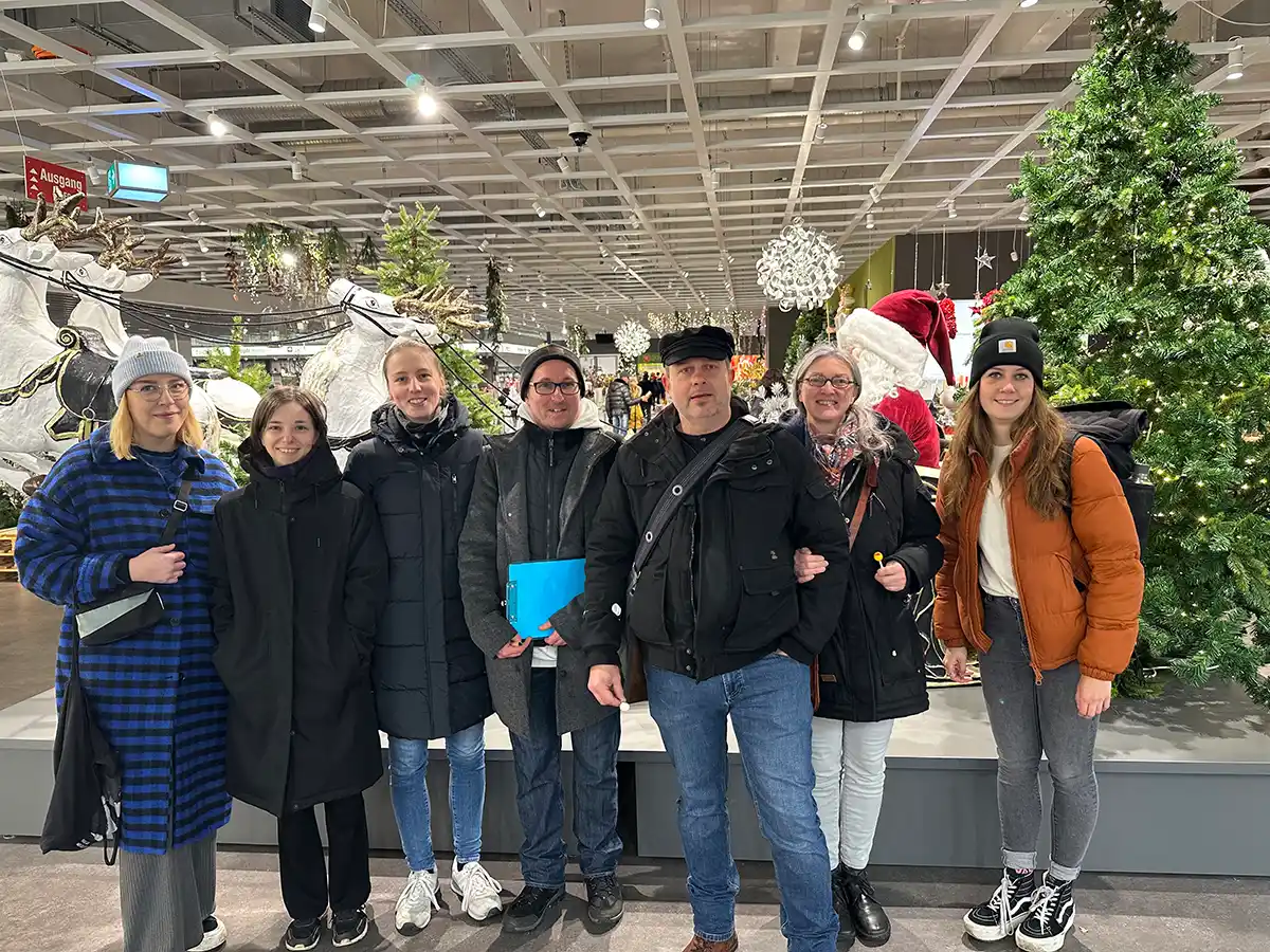 Sieben Personen in Winterkleidung stehen in einem Innenraum in der Nähe von Weihnachtsschmuck, darunter eine große Weihnachtsmannfigur und ein künstlicher Baum.