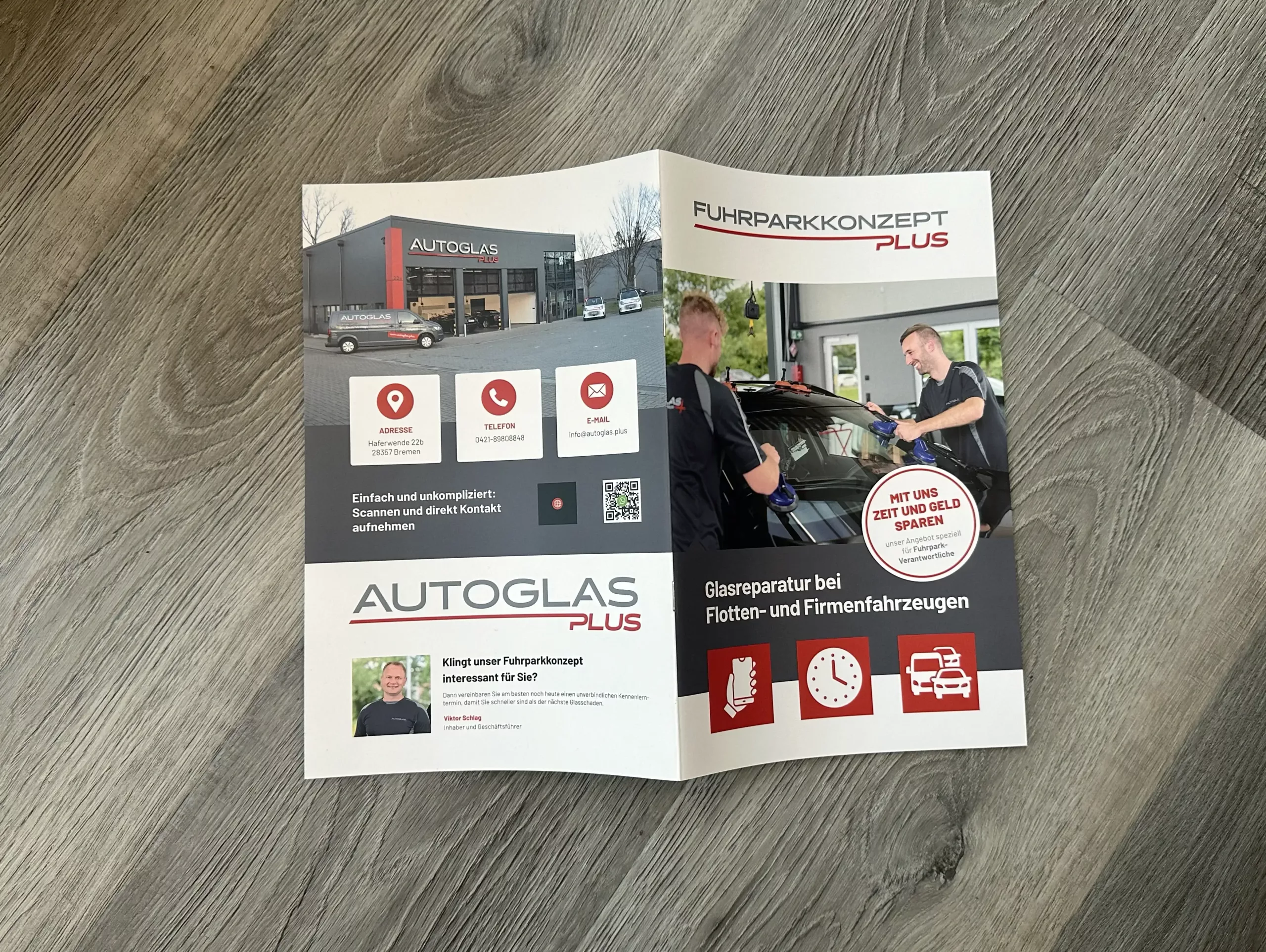 Eine Broschüre für "Autoglas Plus" und "Fuhrparkkonzept Plus" liegt aufgeschlagen auf einem Holzboden, mit Bildern, Kontaktsymbolen und Informationen über Autoglas-Dienstleistungen für Fuhrparks.