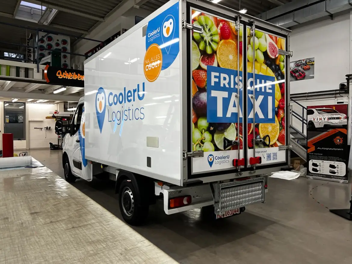 Ein weißer Lieferwagen von CoolerU Logistics mit bunten Obstmotiven und "FRISCHE TAXI" auf der Rückseite, der in einer Garage oder einem Lagerhaus geparkt ist.
