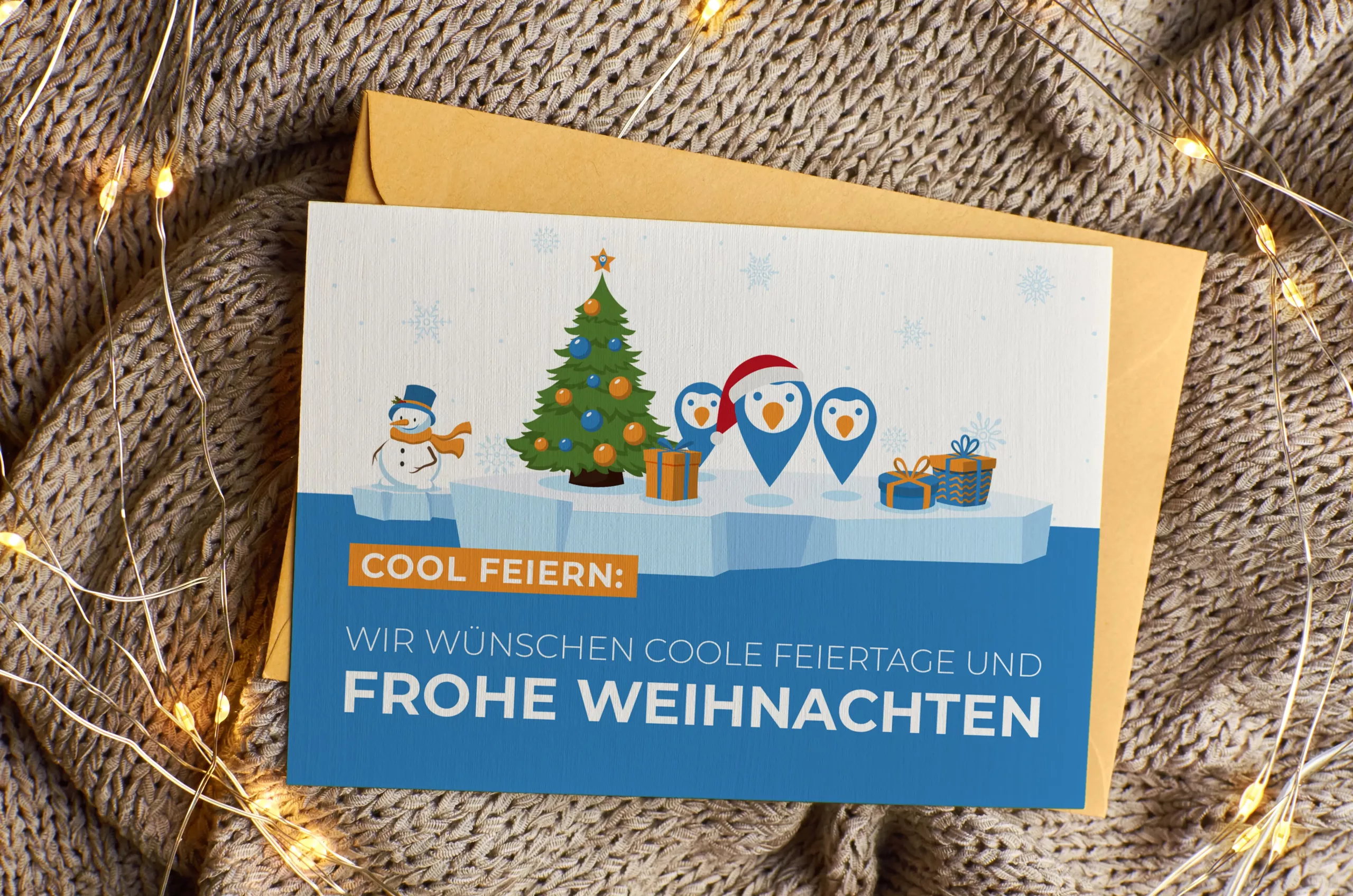 Eine Weihnachtskarte mit Pinguinen, einem Weihnachtsbaum und Geschenken auf einer Eisscholle, daneben ein Text auf Deutsch, der kühle Feiertage und frohe Weihnachten wünscht, platziert auf einer Strickdecke mit Lichterketten.