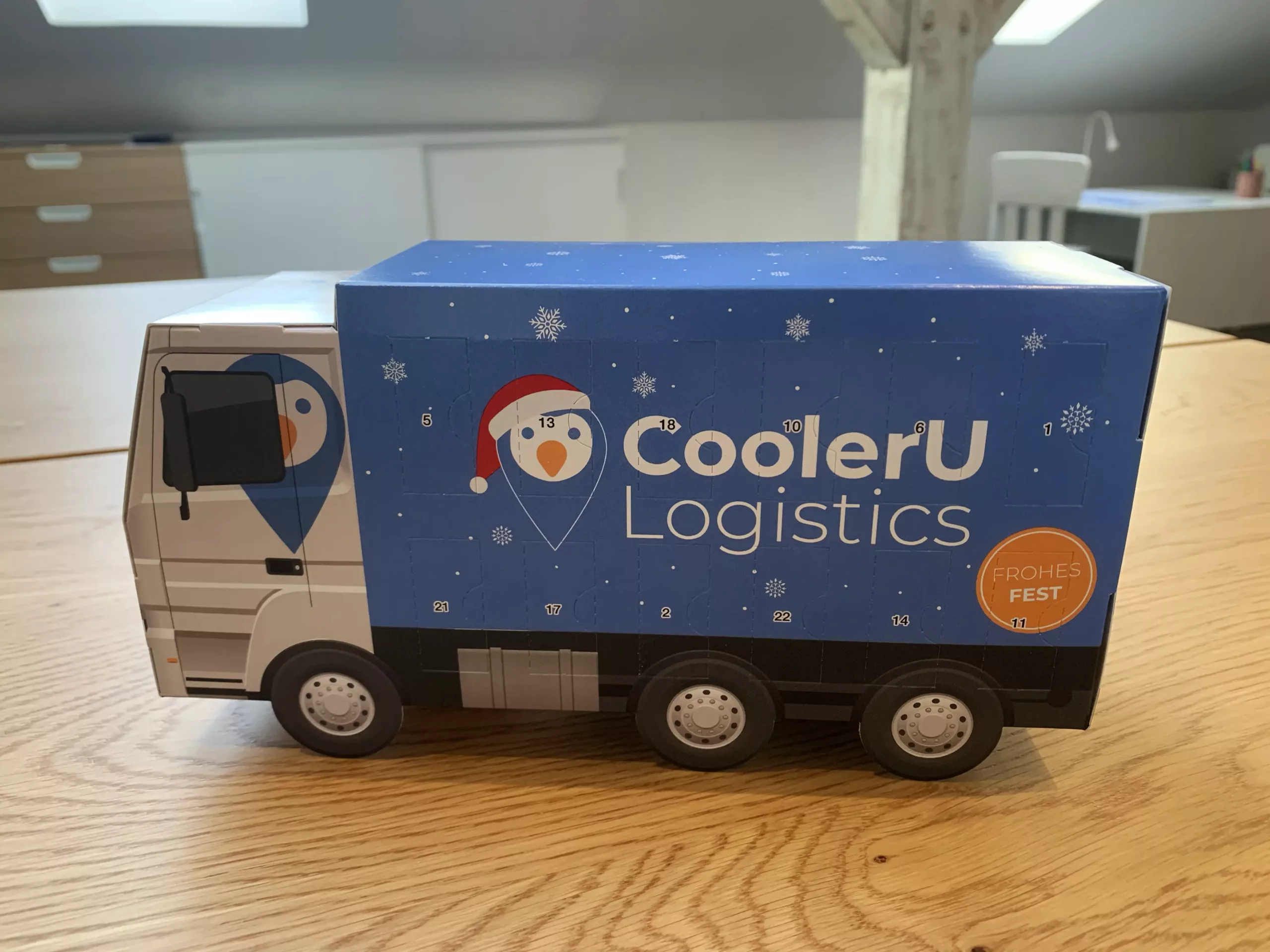 Ein Spielzeug-Lieferwagen mit dem CoolerU Logistics"-Schriftzug und einem festlichen Pinguin-Motiv steht auf einem Holztisch in einer modernen Büroumgebung.