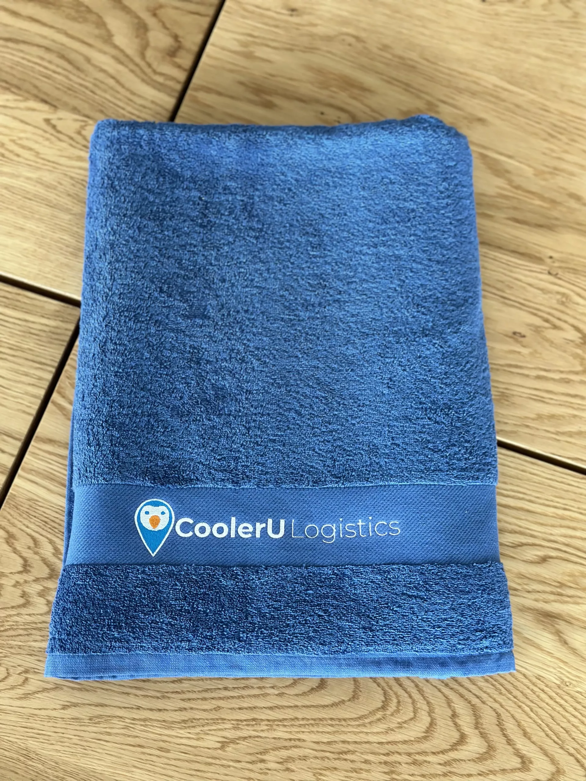 Ein gefaltetes blaues Handtuch auf einer hölzernen Oberfläche mit einem CoolerU Logistics Markenband umwickelt.