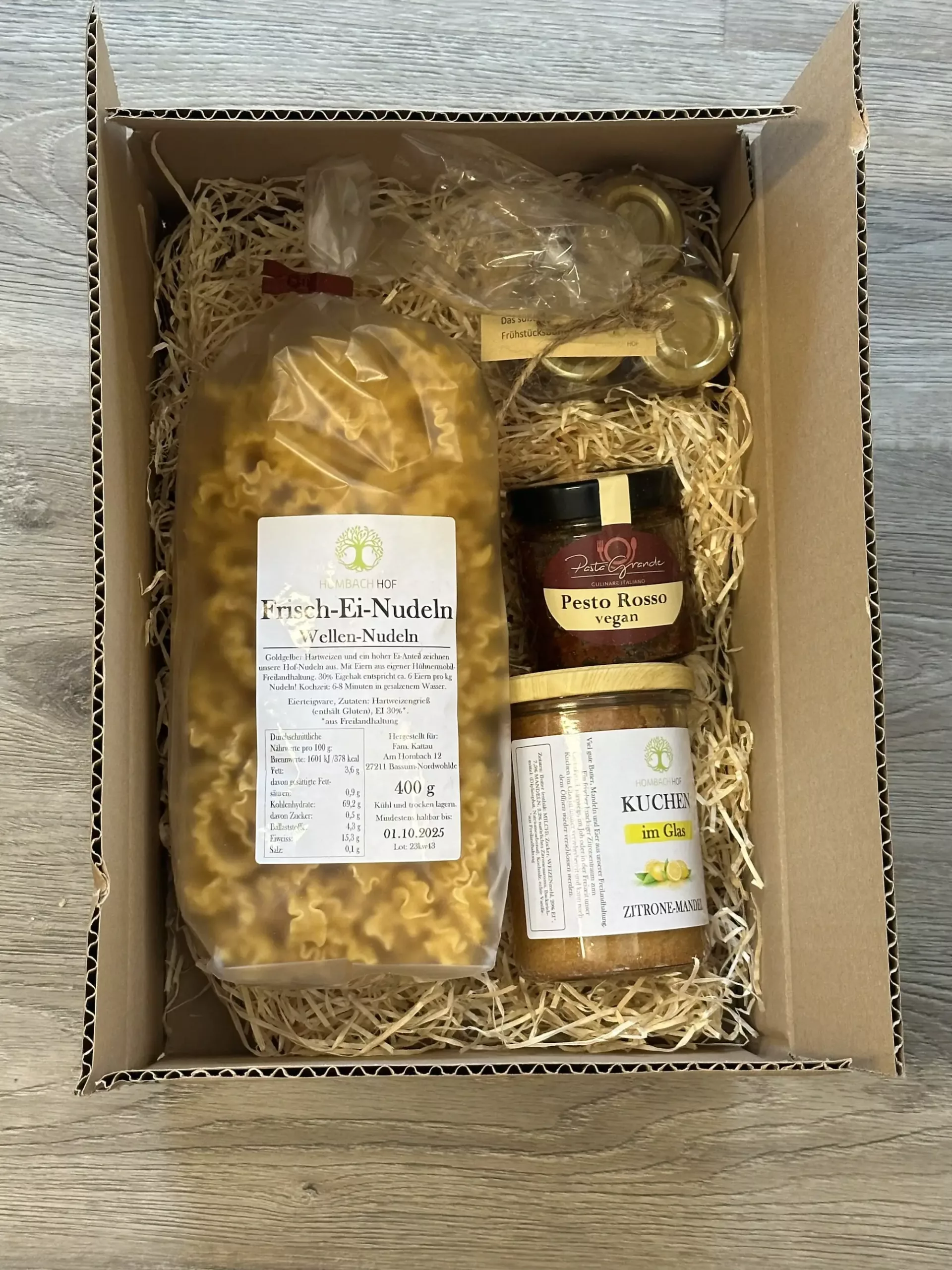 Eine Geschenkpackung mit einer Tüte gewellter Eiernudeln, einem Glas veganem roten Pesto, einem Glas Zitronensalz und einem kleinen, in Plastik eingewickelten Päckchen auf geschreddertem Papier.