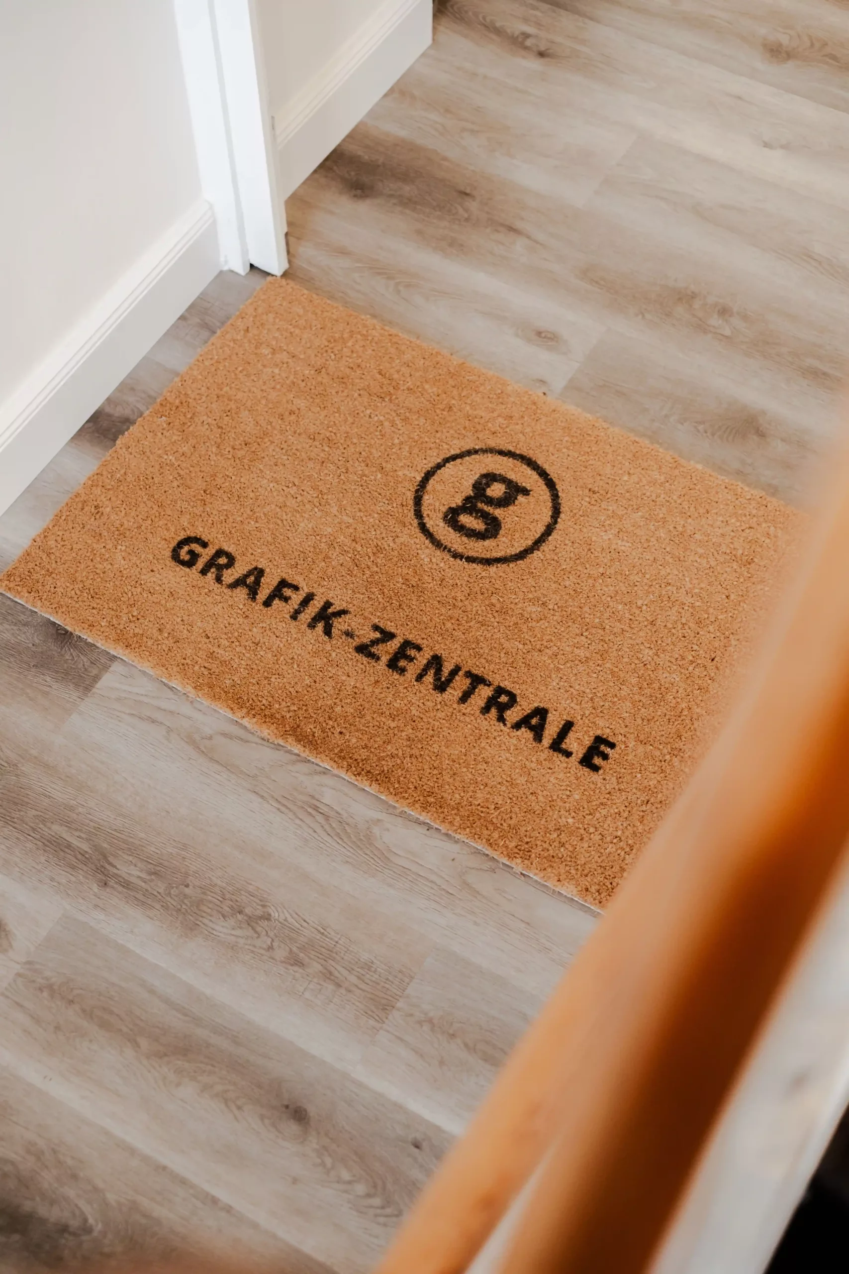 Eine braune Fußmatte mit dem schwarzen Schriftzug "GRAFIK-ZENTRALE" und einem kreisförmigen Logo liegt auf einem hellen Holzboden neben einer weißen Wand.