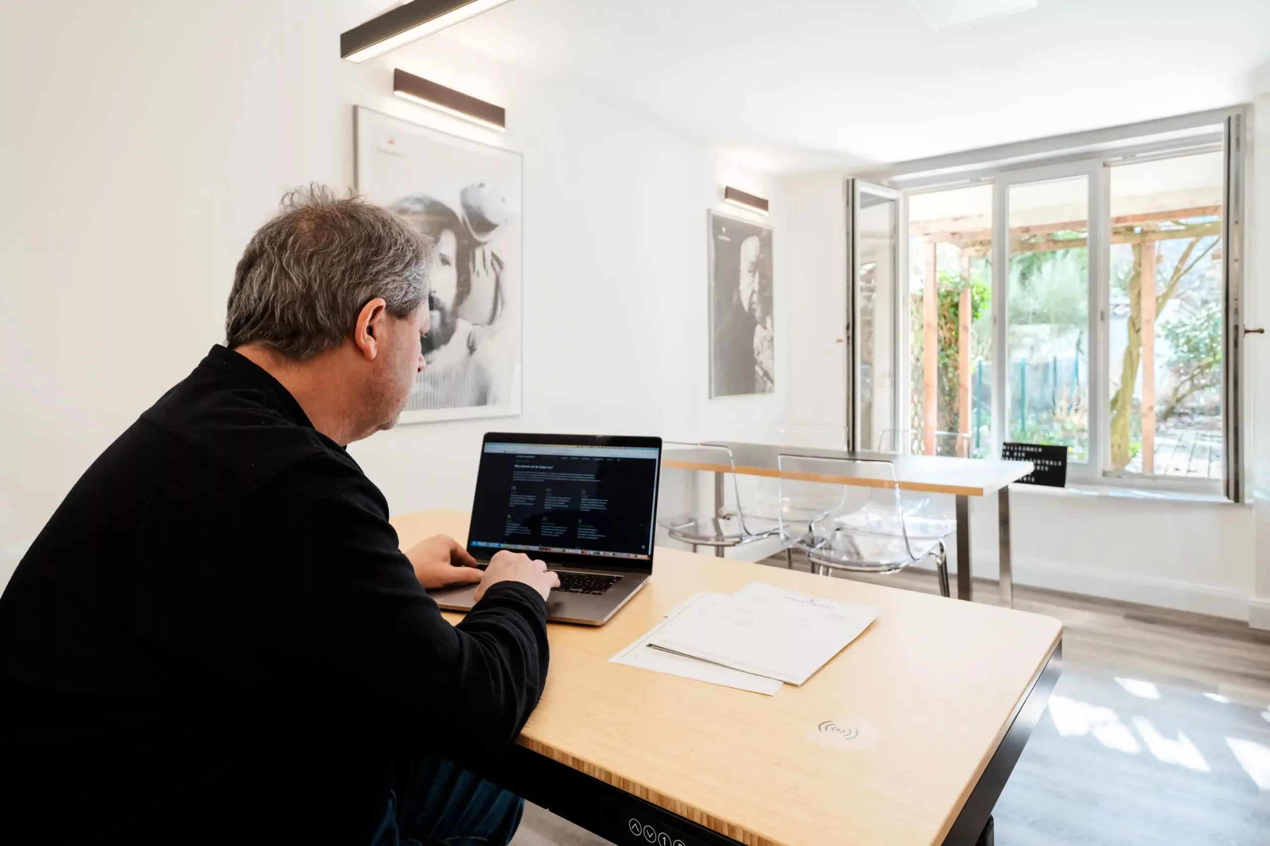 Ein Mann sitzt an einem Schreibtisch und arbeitet an einem Laptop in einem hellen, modernen Büro mit Papieren in der Nähe und einem offenen Fenster im Hintergrund.