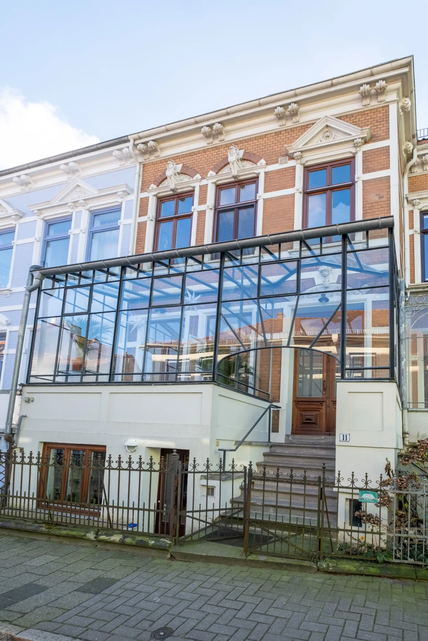 Ein Backstein-Stadthaus mit großen Fenstern und einer modernen, verglasten Veranda über einem Eingangstor und einer Treppe, die zur Eingangstür führt.