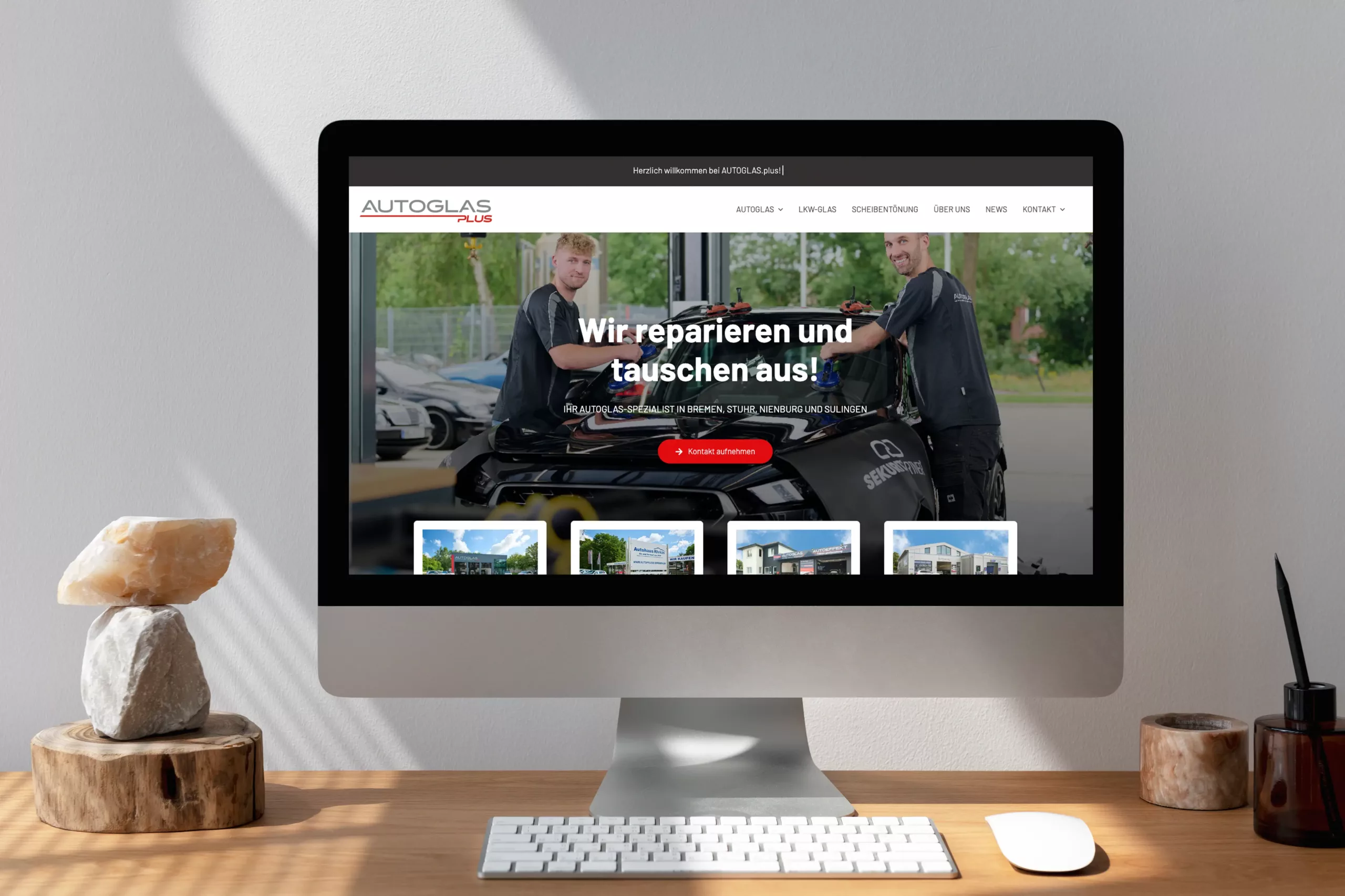 Computerbildschirm mit der Website von Autoglas Plus, auf dem zwei Arbeiter bei der Reparatur einer Windschutzscheibe zu sehen sind, mit Text in deutscher Sprache und sichtbaren Navigationsmenüs.