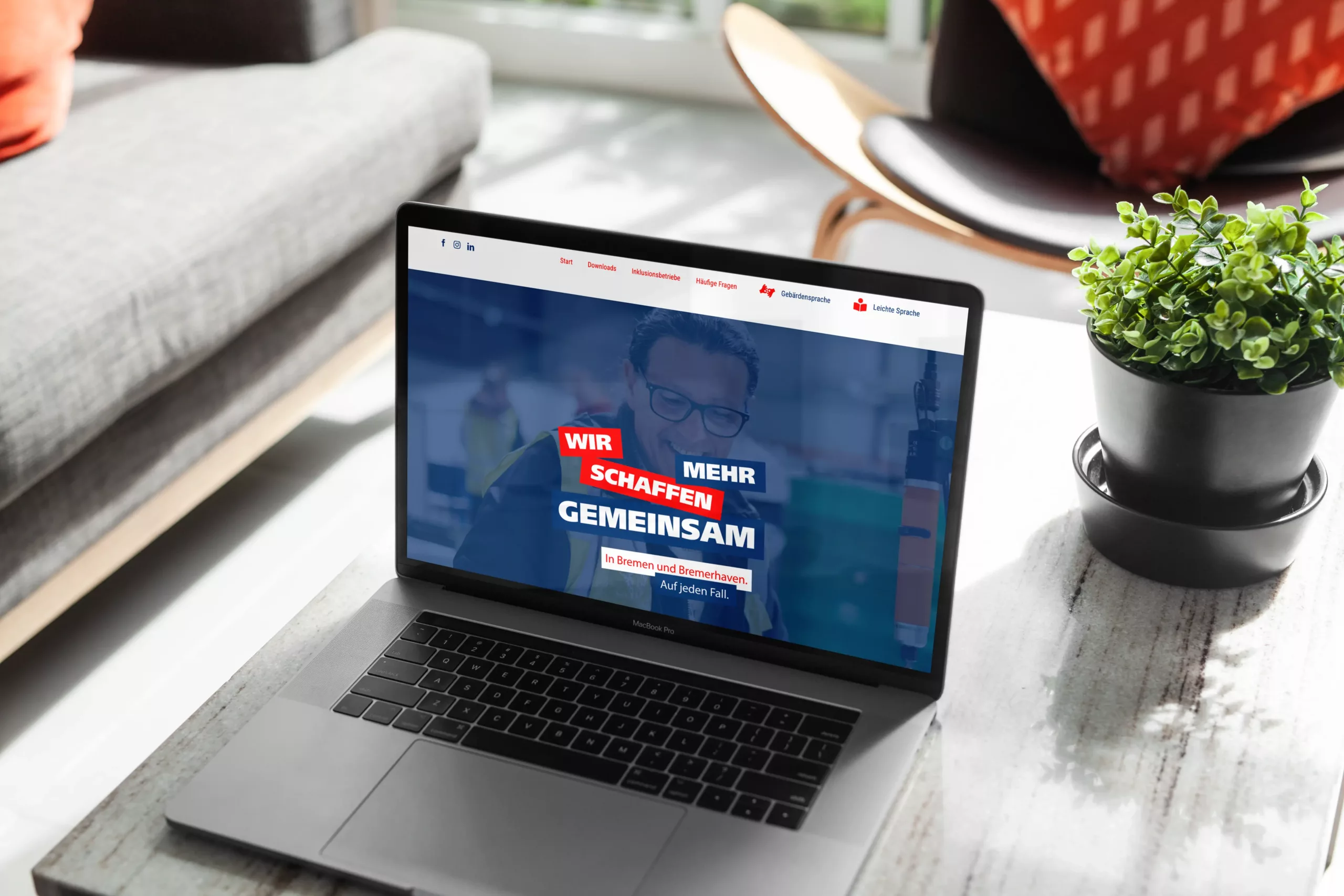 Ein Laptop auf einem Couchtisch zeigt eine Website mit dem deutschen Text "Wir schaffen mehr gemeinsam" in einem modernen Wohnzimmer mit einer Pflanze und einem Sofa im Hintergrund.
