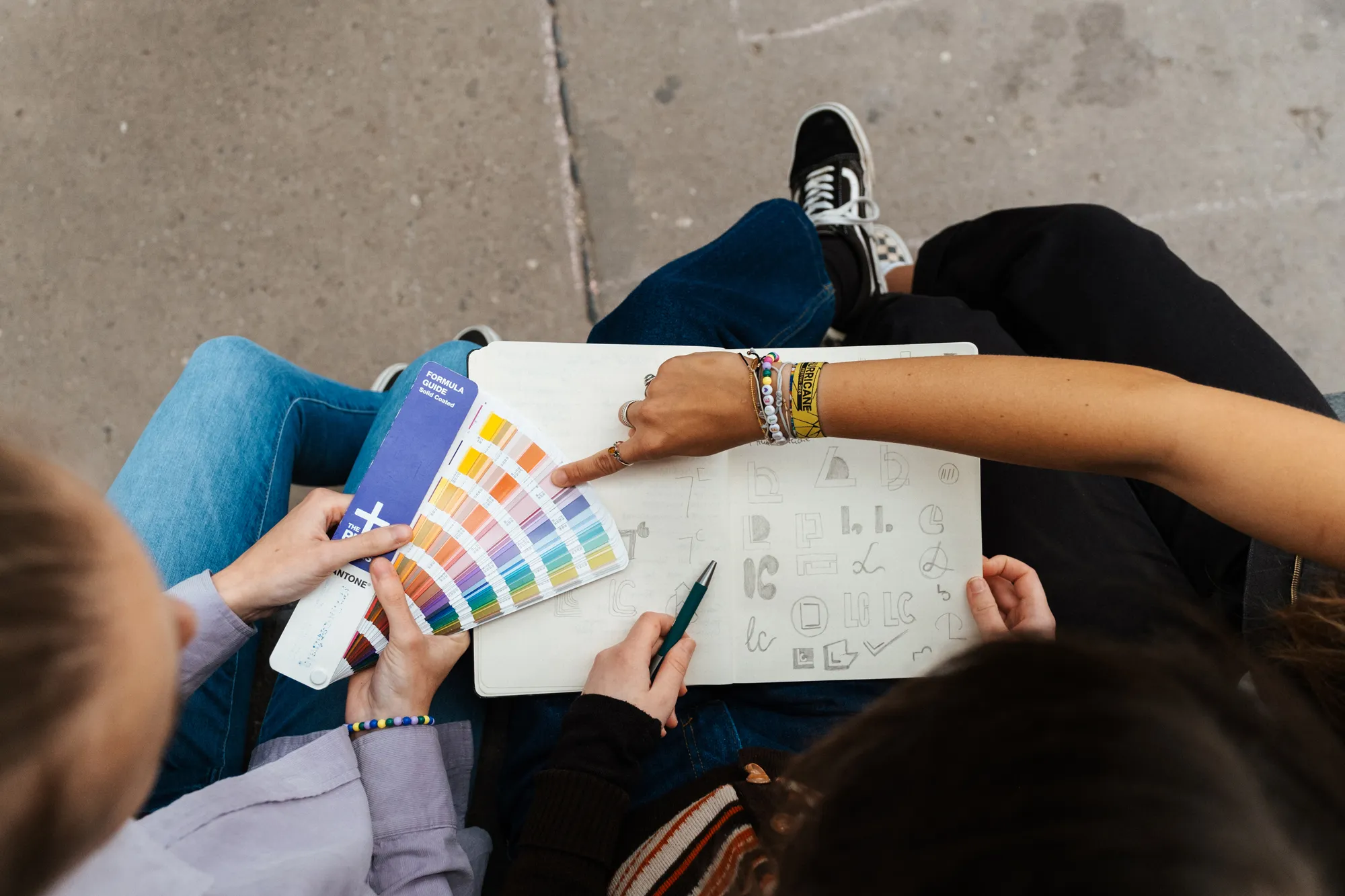 grafik-zentrale-werbeagentur-bremen-unsere-arbeit Drei Personen sitzen zusammen, eine hält eine Farbpalette in der Hand, eine andere zeigt auf ein Skizzenbuch, das mit handgezeichneten Buchstaben und Formen gefüllt ist, während die dritte Person einen Stift hält.