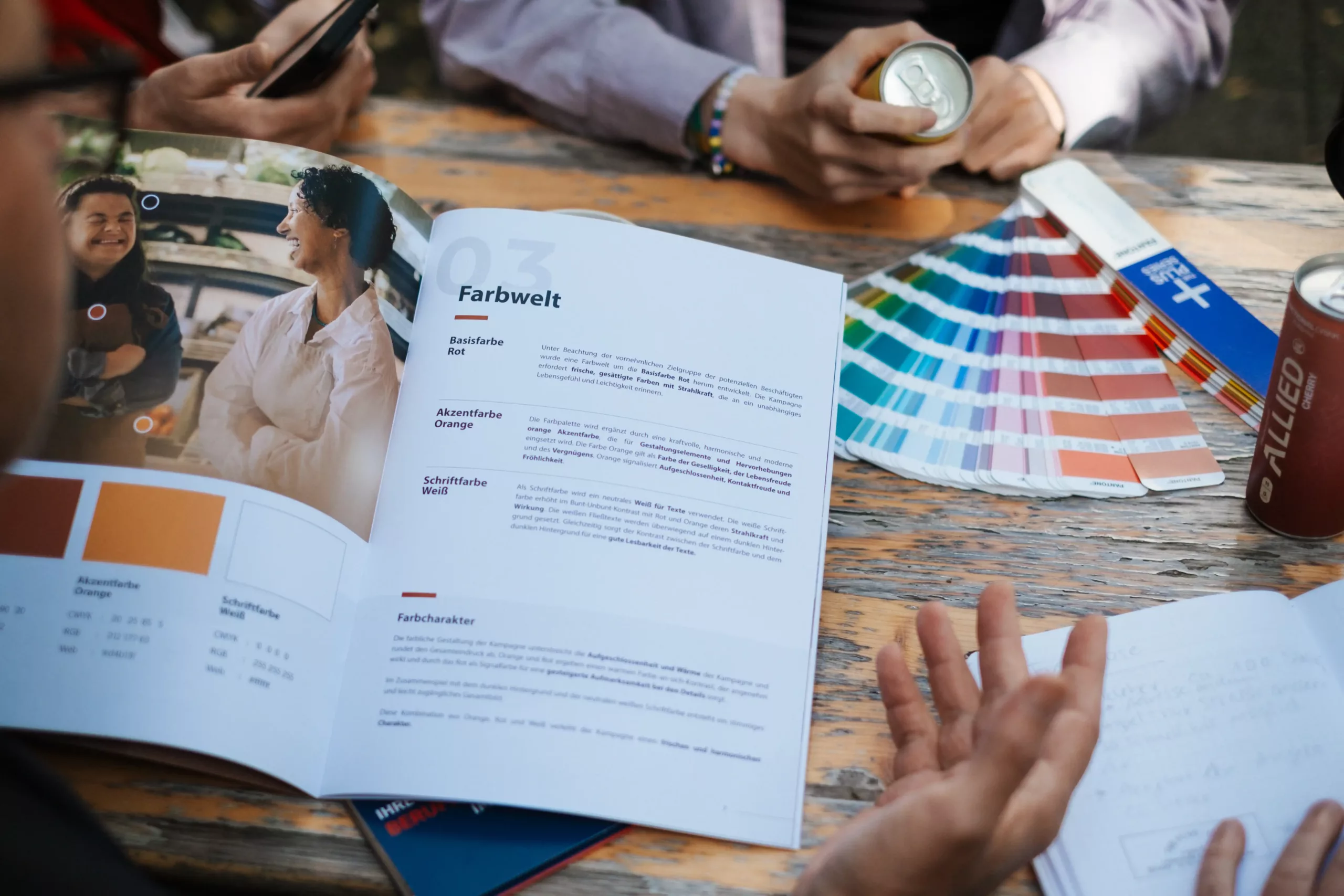 grafik-zentrale_werbeagentur_bremen_Corporate-Design_Entwicklung_Design_Manual Eine Gruppe von Personen diskutiert an einem Holztisch mit einem aufgeschlagenen Farbkatalog, Farbmustern, Getränken und Notizbüchern über Farboptionen.