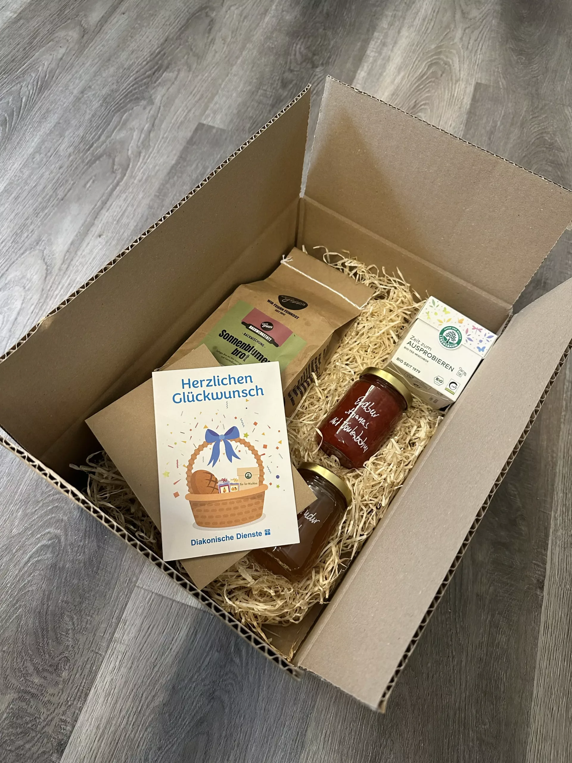 Eine Pappschachtel mit einer Strohverpackung enthält eine Grußkarte, Kaffee, Tee, ein Glas Marmelade und ein Glas Honig und steht auf einem Holzboden.