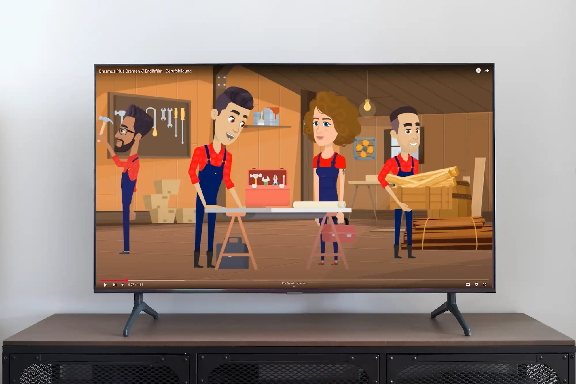 Ein Flachbildschirm zeigt ein animiertes Video von vier Personen in einer Werkstatt, die rotkarierte Hemden und Overalls tragen und mit Holz und Werkzeug arbeiten.