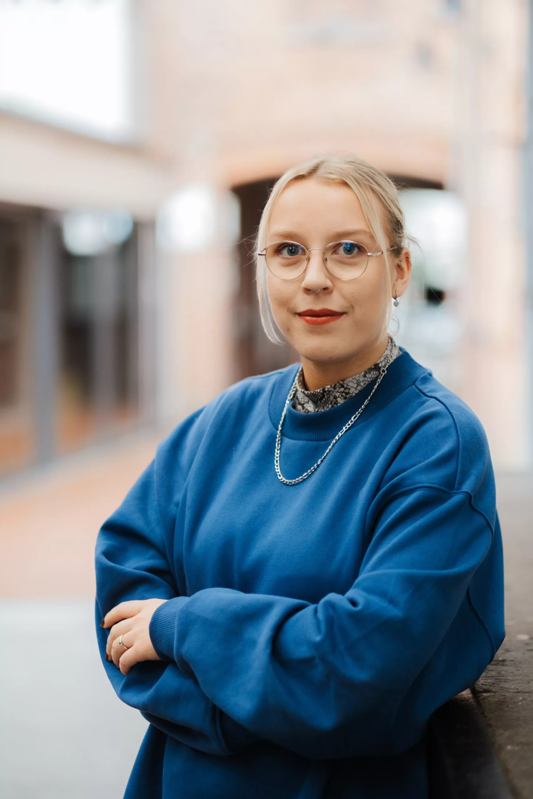 Eine Person mit blondem Haar, Brille und rotem Lippenstift trägt ein blaues Sweatshirt und steht mit verschränkten Armen in einer städtischen Umgebung.