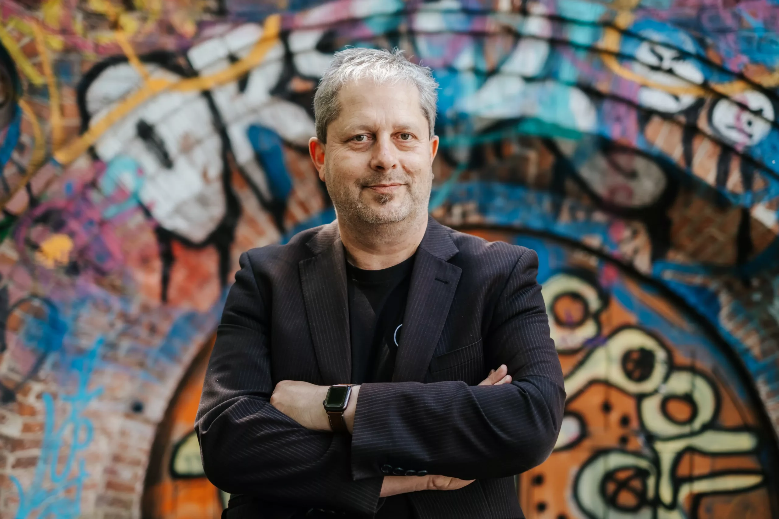 Ein Mann in einem dunklen Blazer steht mit verschränkten Armen vor einer bunten, mit Graffiti beschmierten Ziegelwand.