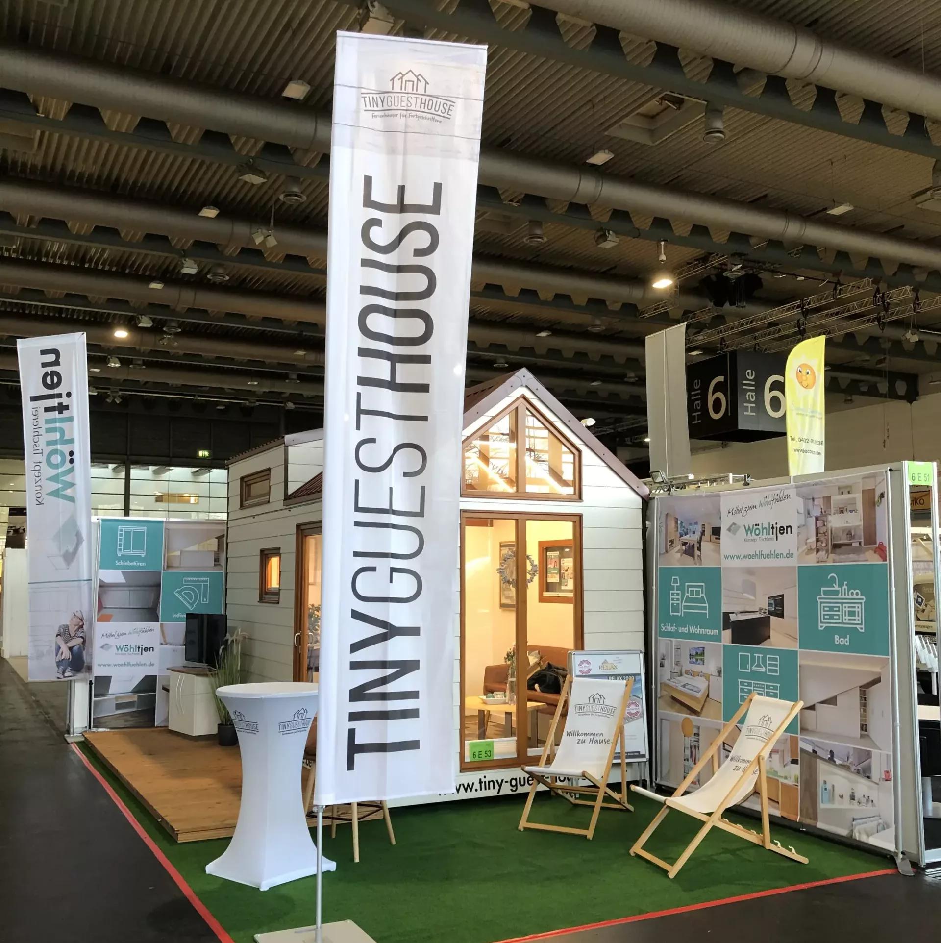 Ein Ausstellungsstand für ein winziges Haus auf einer Indoor-Messe, mit einem weißen Modell eines winzigen Hauses, Informationsbannern und Liegestühlen auf Kunstrasen.