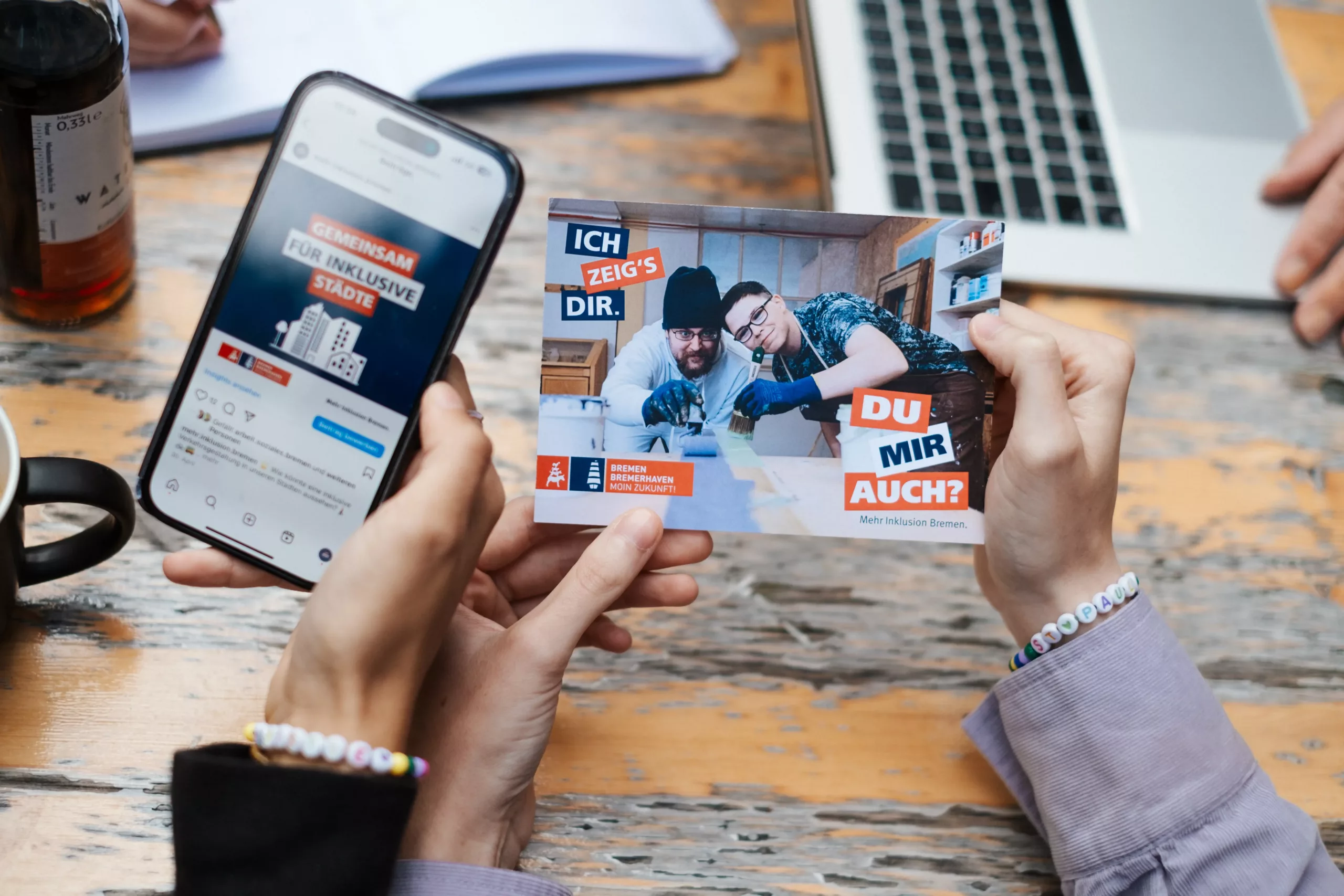Zwei Personen halten ein Smartphone und eine Postkarte in der Hand. Die Postkarte zeigt zwei Männer bei der Arbeit, mit fettgedrucktem deutschen Text und orangefarbenen, blauen und weißen Designelementen.