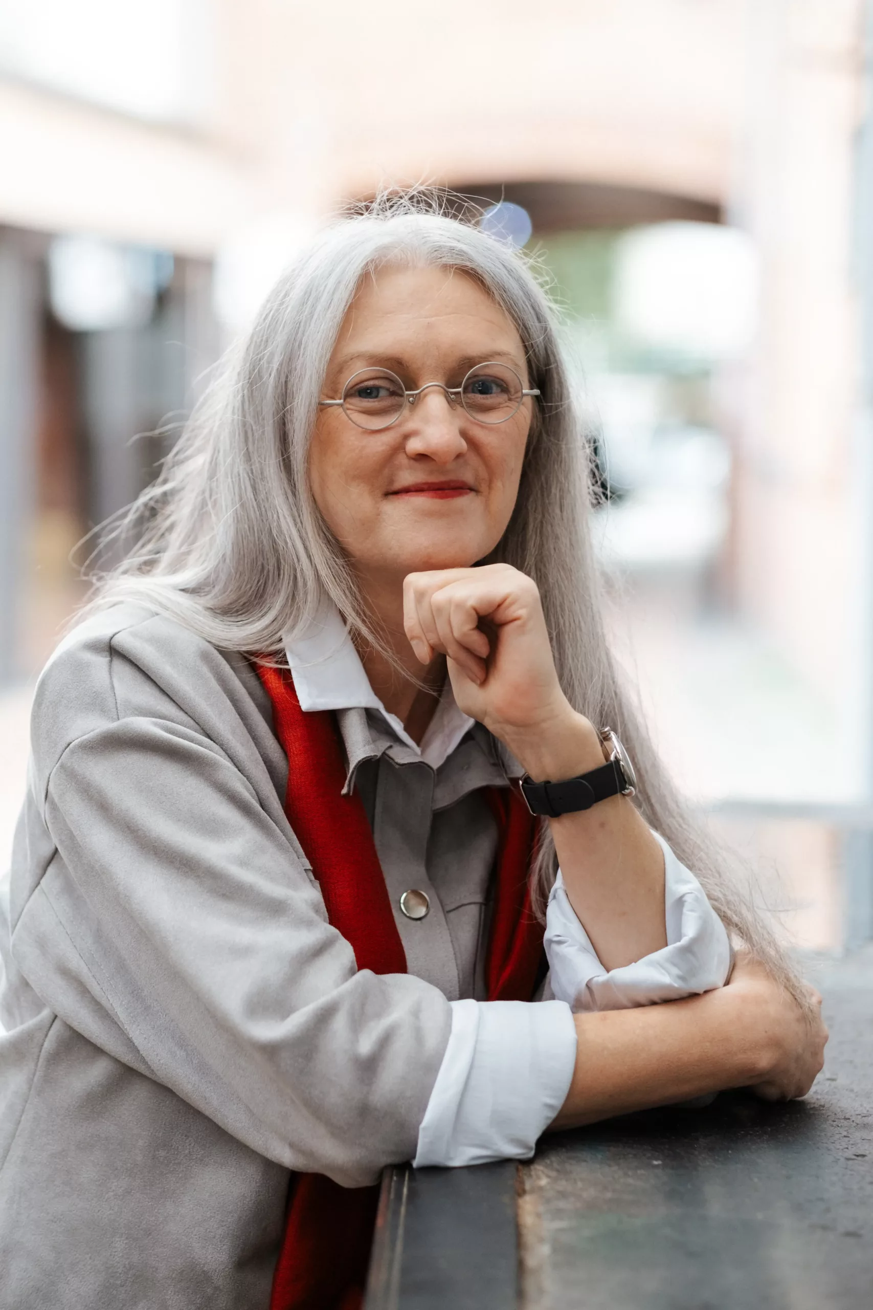 Eine Frau mit langen grauen Haaren, Brille und rotem Schal sitzt an einem Tisch im Freien, stützt ihr Kinn auf die Hand und schaut in die Kamera.