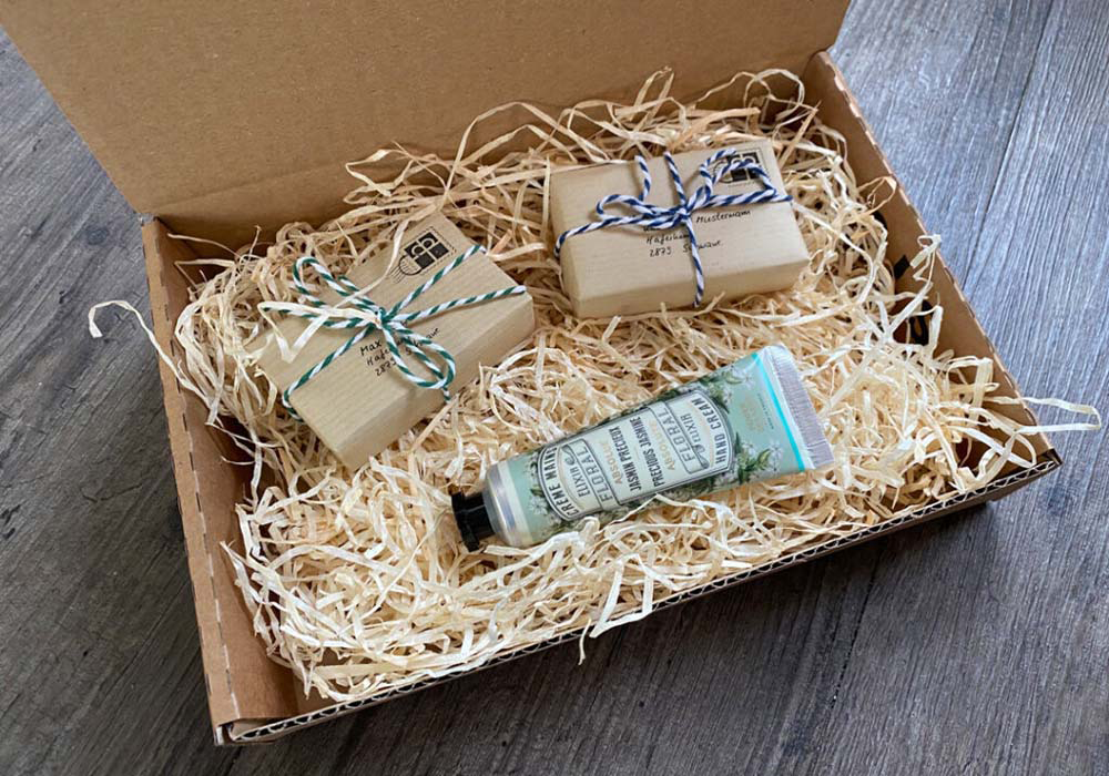 Ein offener Karton, gefüllt mit geschreddertem Verpackungsmaterial, enthält zwei verpackte Seifenstücke und eine Tube Handcreme.