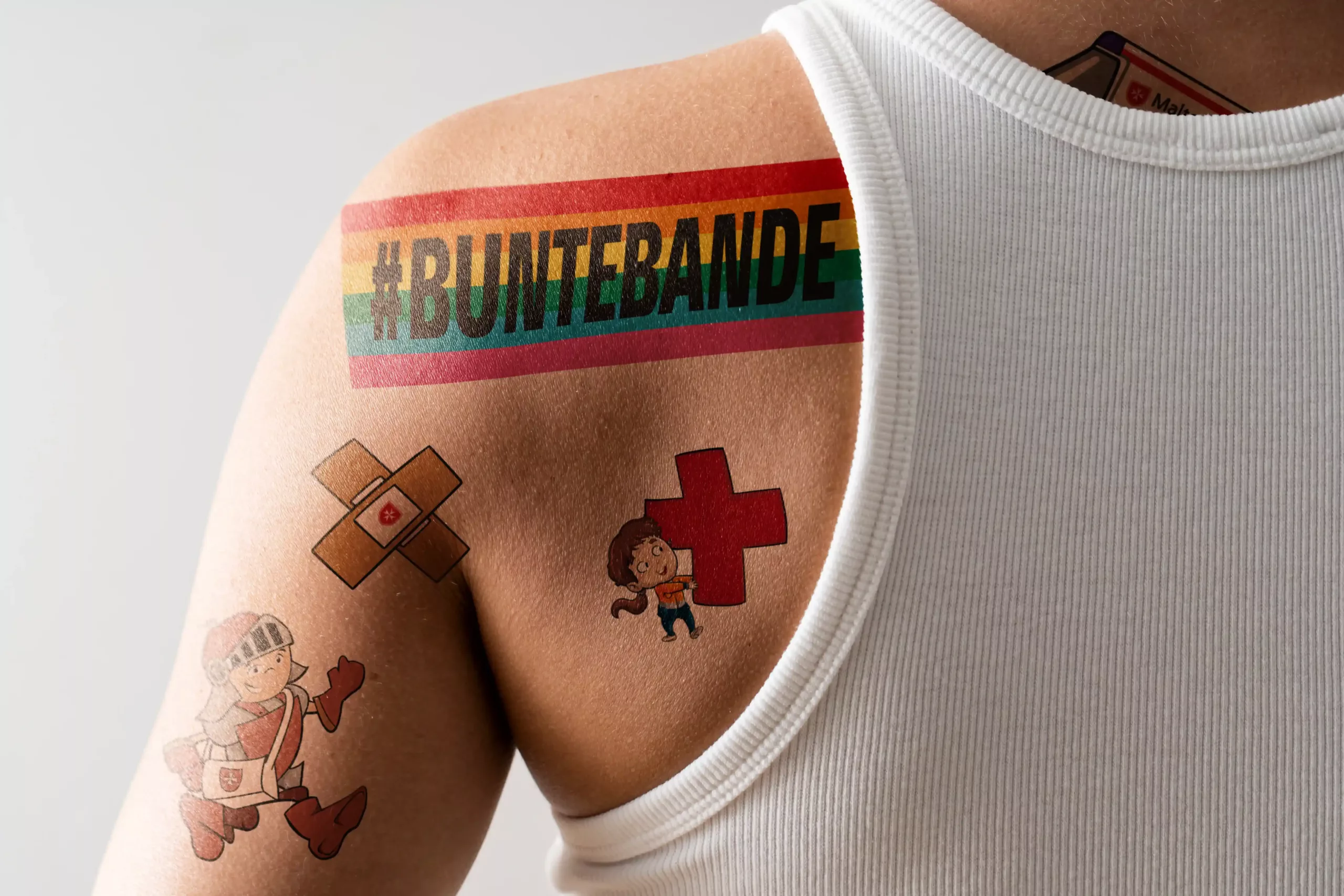 Eine Person in einem weißen Tank-Top mit bunten Tattoos auf der Schulter, darunter "#BUNTEBANDE", ein rotes Kreuz, Bandagen und Cartoon-Figuren.