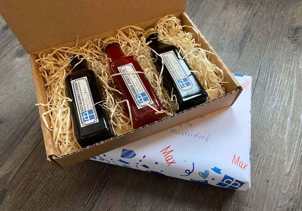 Eine offene Schachtel mit drei Glasflaschen, die mit verschiedenfarbigen Flüssigkeiten auf Strohhalm gefüllt sind, die auf einem teilweise sichtbaren, eingepackten Geschenk liegen.