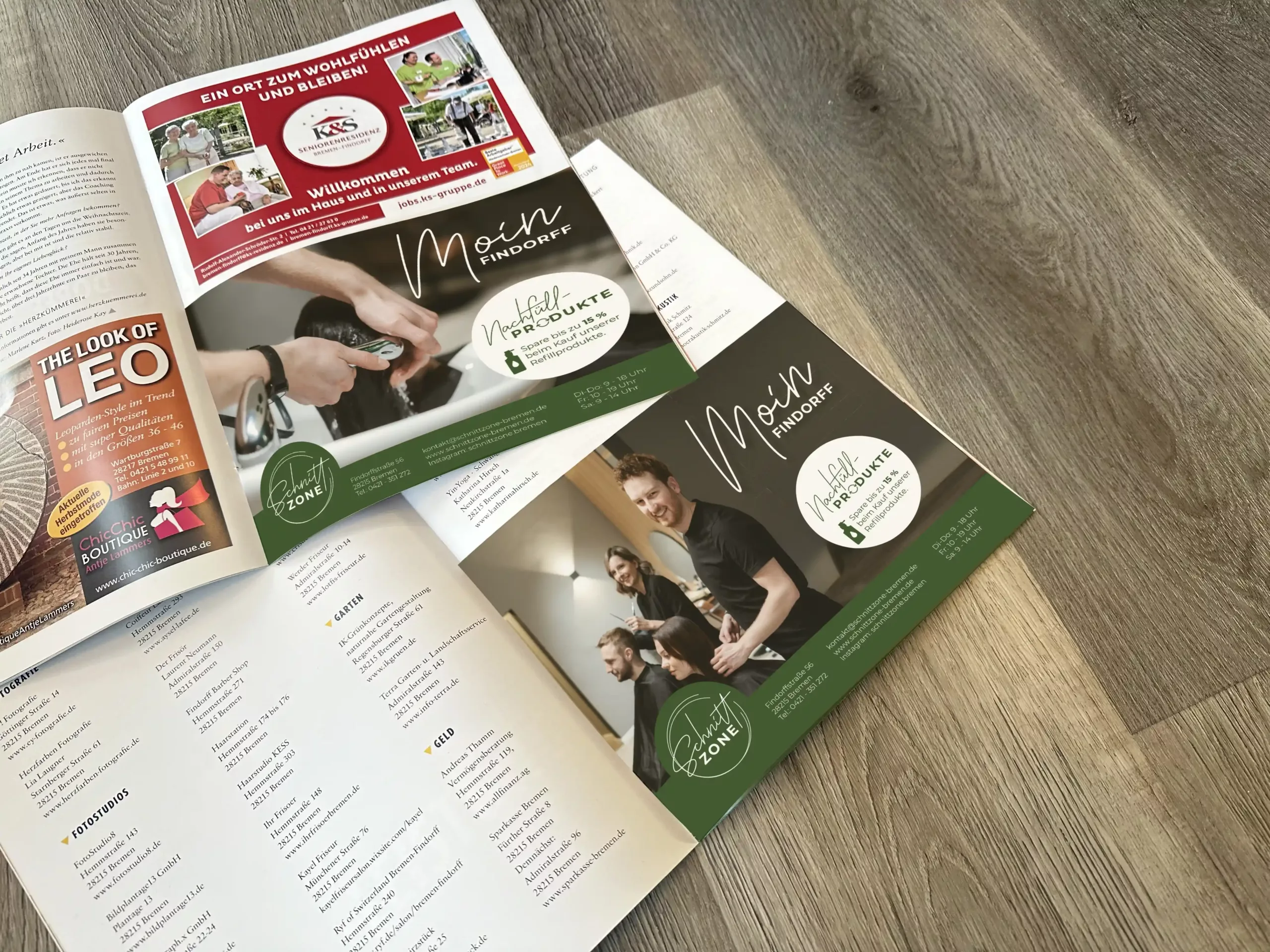 Aufgeschlagene Zeitschriften auf einem Holzboden zeigen Seiten mit Werbung und Artikeln, darunter Bilder von Menschen in einem Friseursalon und verschiedene Texte und Grafiken.