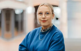Eine Person mit blondem Haar, Brille und blauem Sweatshirt steht in einem Haus und schaut mit neutralem Blick in die Kamera.