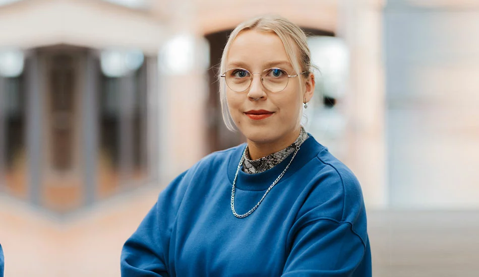 Eine Person mit blondem Haar, Brille und blauem Sweatshirt steht in einem Haus und schaut mit neutralem Blick in die Kamera.