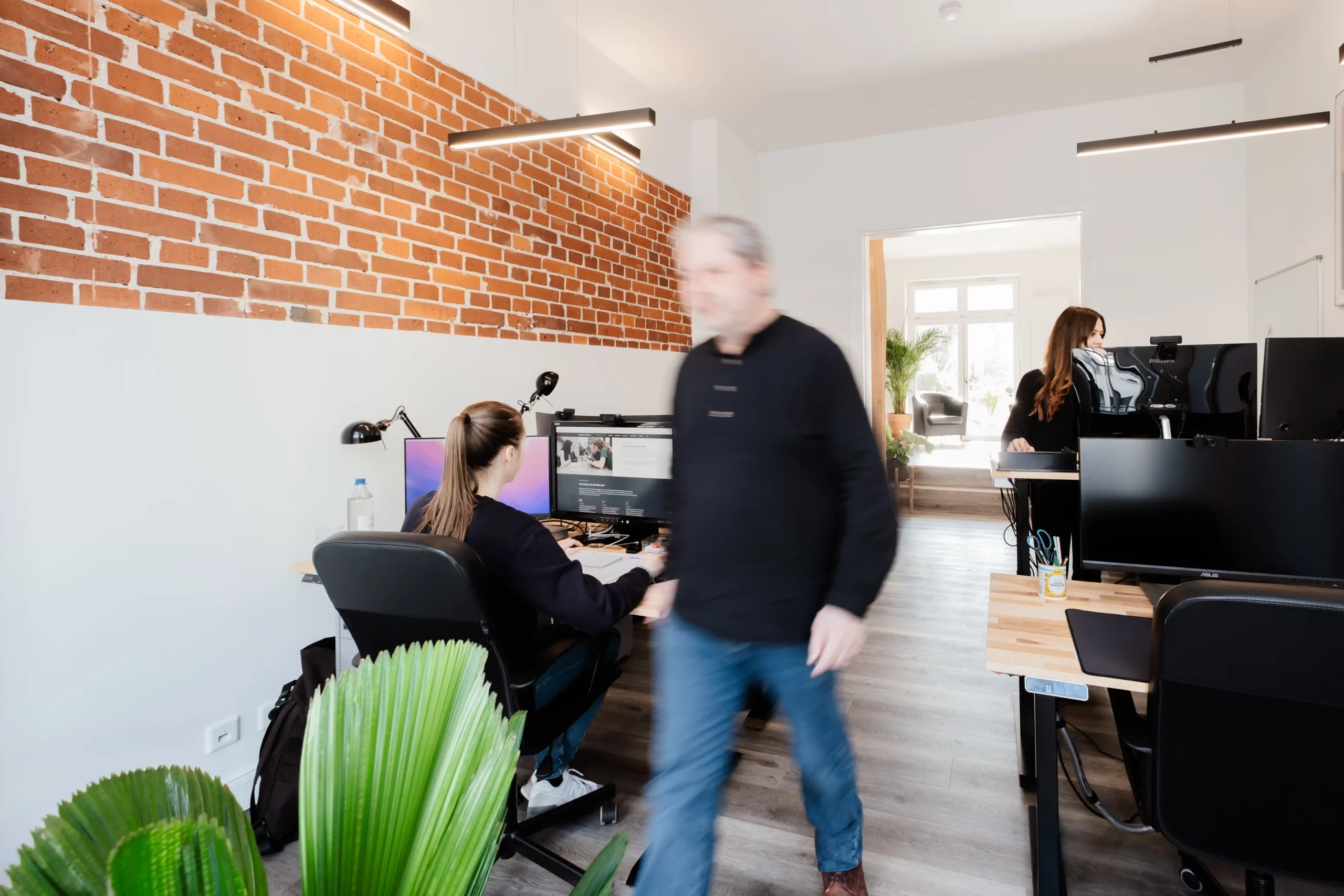 Modernes Büro mit freiliegender Backsteinwand, Menschen, die an Schreibtischen mit Computern arbeiten, und eine Person, die durch den Raum geht.