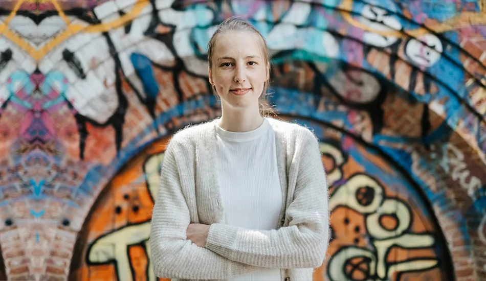 Eine Frau steht mit verschränkten Armen vor einer mit bunten Graffiti bedeckten Backsteinmauer.