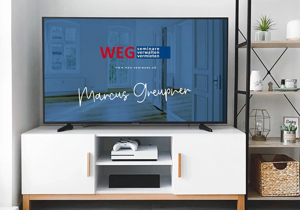 Flachbildfernseher auf einem weißen TV-Schrank, der eine Website mit dem Text "WEG - seminare verwalten vermieten Marcus Greupner" zeigt. Eine Spielkonsole und Dekorationsartikel sind ebenfalls zu sehen.