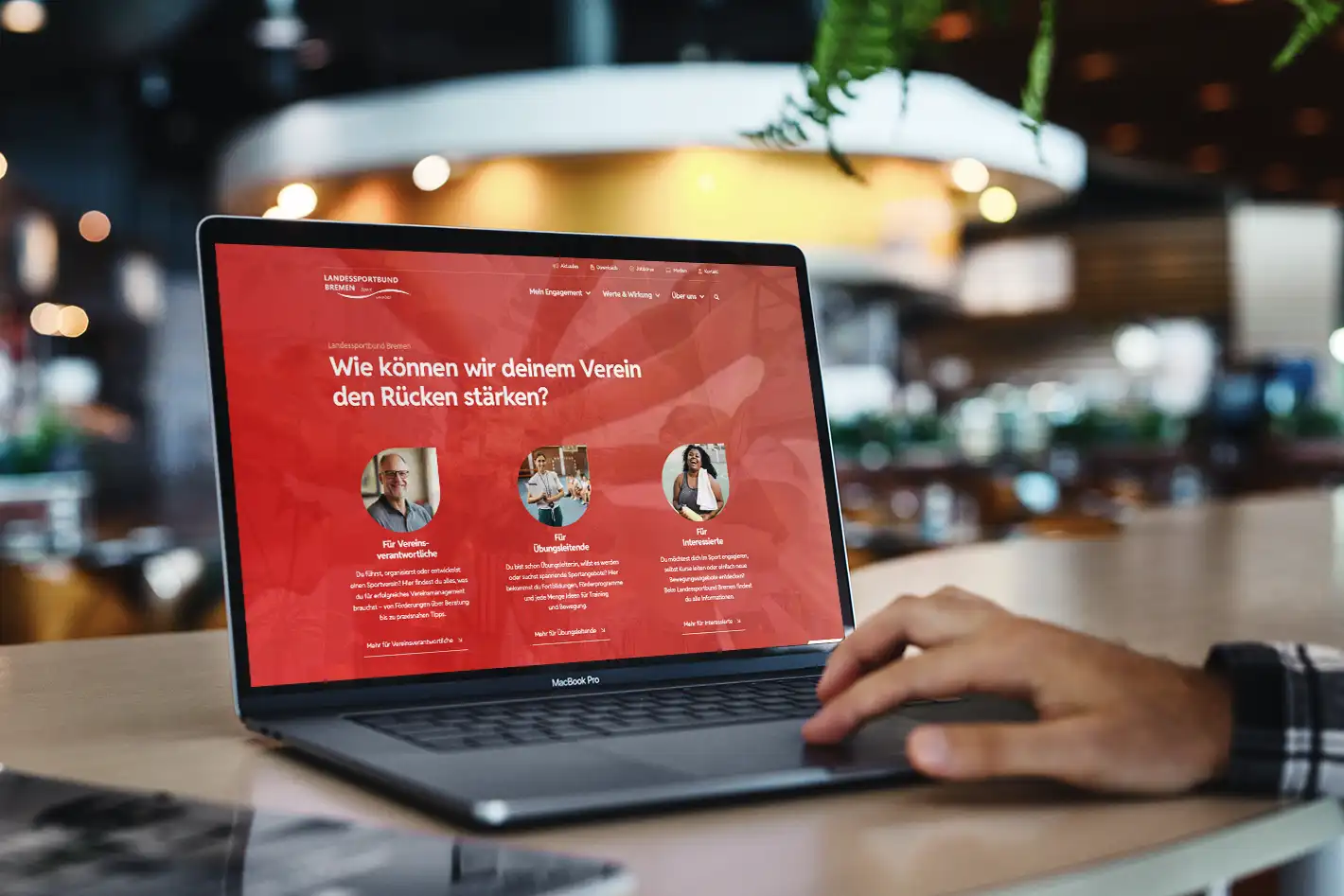 Eine Person benutzt ein MacBook Pro, das eine Website mit der Überschrift "Wie können wir deinem Verein den Rücken stärken?" in einem modernen Café anzeigt.