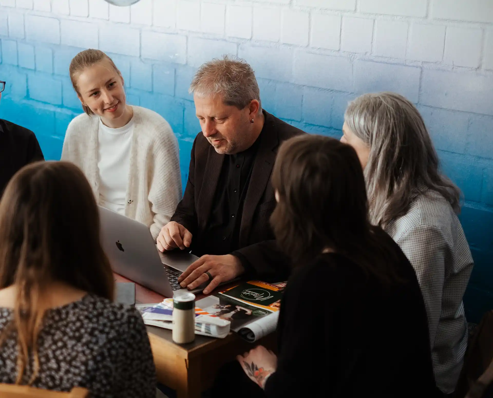 Eine Gruppe von fünf Personen sitzt an einem Tisch, schaut auf einen Laptop und diskutiert vor einem blau-weißen Backsteinhintergrund.