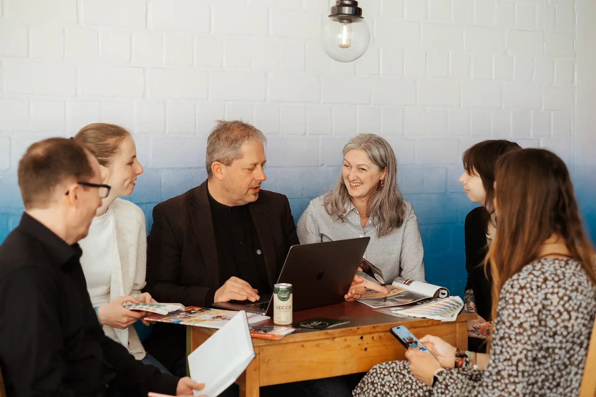 Sechs Personen sitzen um einen kleinen Tisch mit einem Laptop, Zeitschriften und Getränken und diskutieren in einer lockeren, gut beleuchteten Umgebung.