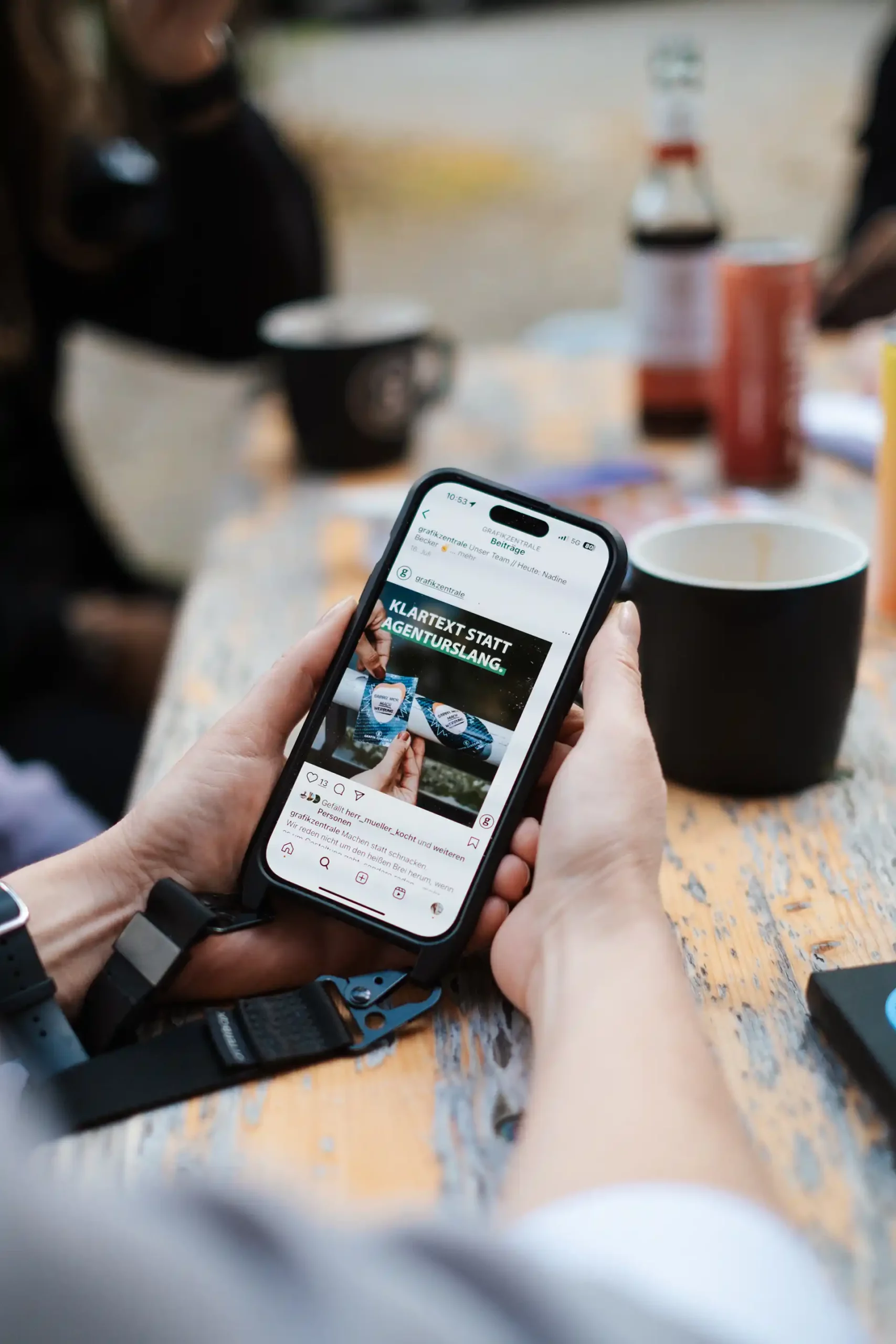 Eine Person hält ein Smartphone an einem Tisch im Freien und surft auf einer Website; auf dem Holztisch stehen Kaffeetassen und Flaschen.