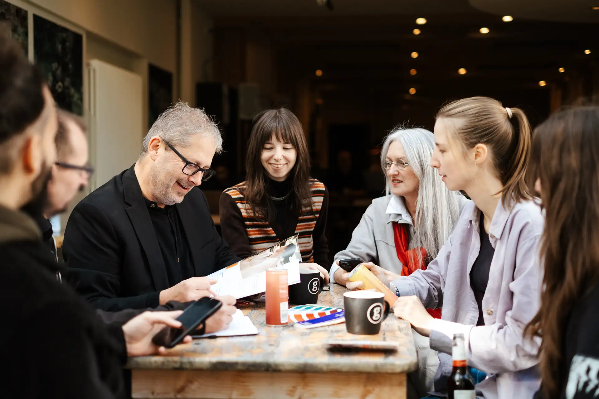 Werbeagentur_Bremen_grafik-zentrale_Workshop_team Eine Gruppe von Menschen sitzt um einen Holztisch mit Getränken und Papieren und unterhält sich in einem gut beleuchteten Café.