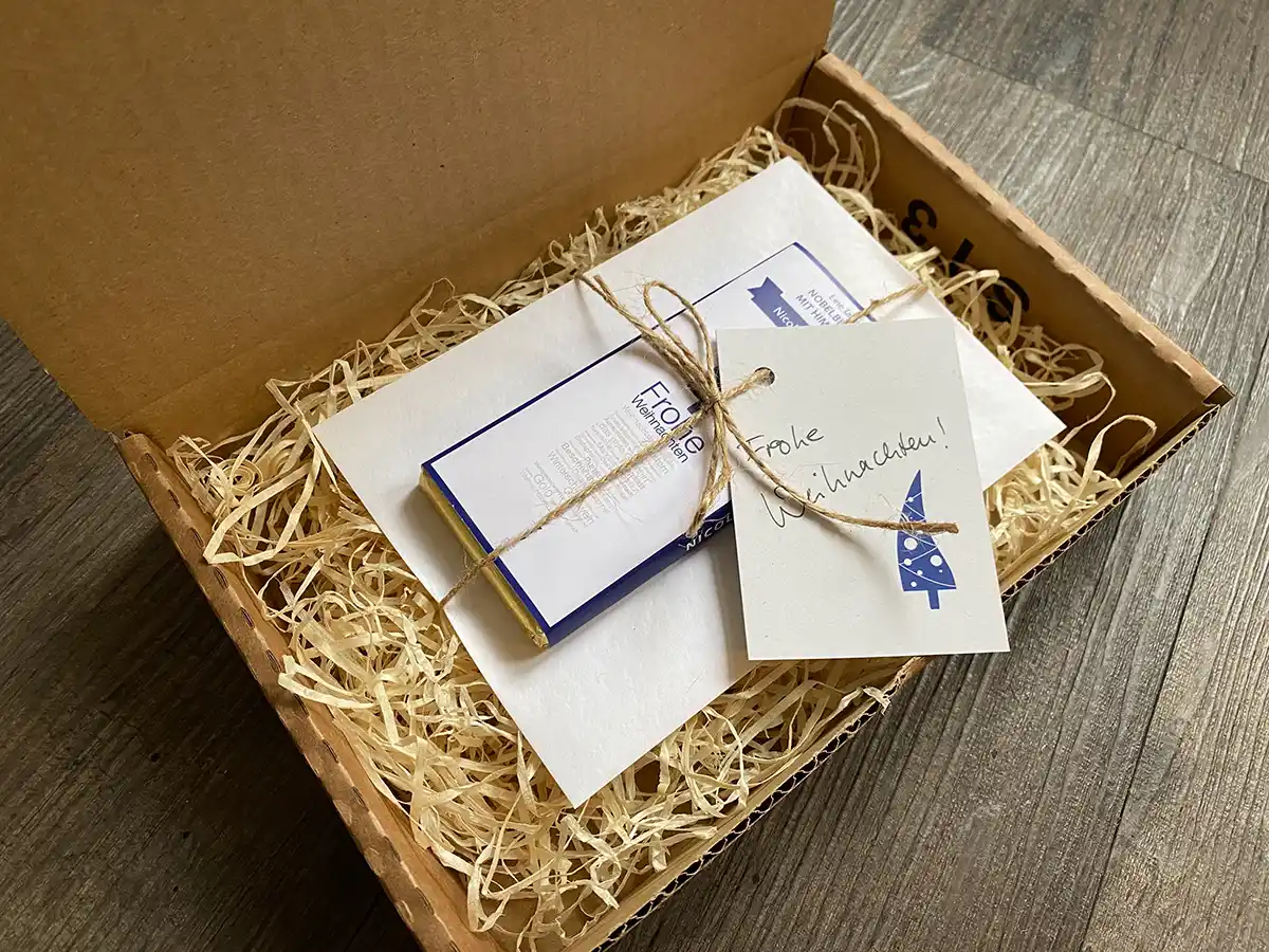 Ein mit geschreddertem Papier gefüllter Karton enthält einen Schokoriegel, einen Umschlag und einen mit Schnur gebundenen Zettel mit der Aufschrift "Frohe Weihnachten!" und einem blauen Baum.