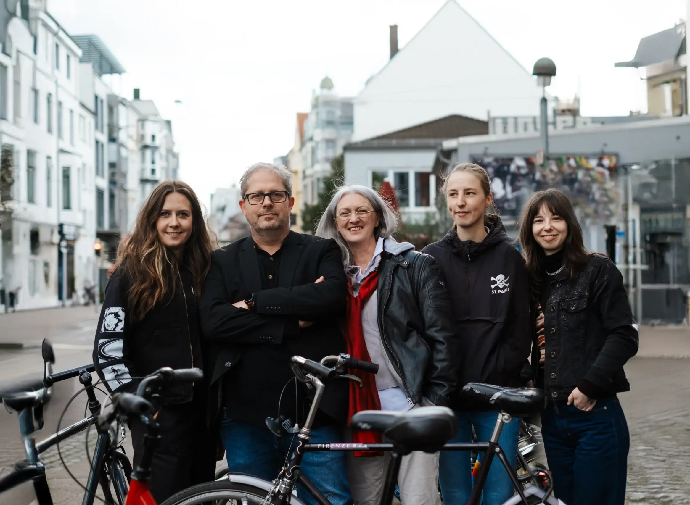 Fünf Personen stehen auf einer Stadtstraße hinter Fahrrädern zusammen und posieren für ein Gruppenfoto. Im Hintergrund sind Gebäude und städtische Elemente zu sehen.