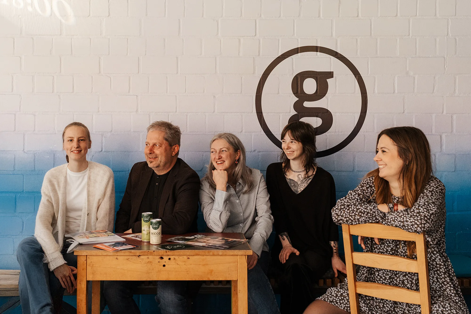 Fünf Personen sitzen an einem Holztisch vor einer weißen Backsteinwand mit einem großen "g"-Logo, lächeln und schauen in verschiedene Richtungen.